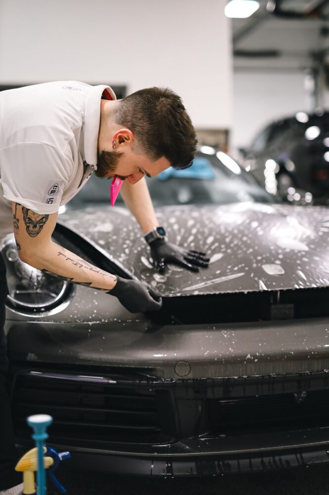 Meistras automobilių dirbtuvėse kruopščiai klijuoja apsauginę plėvelę ant automobilio kėbulo.